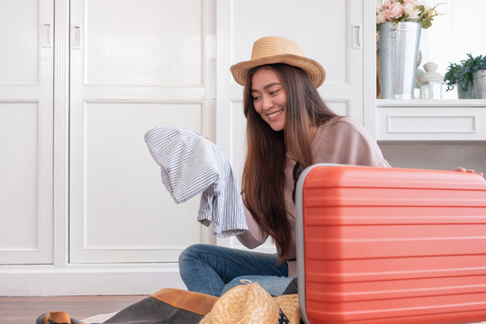 Young asian woman traveler prepare cloth into orange suitcase for holiday vacation at home.backpacker travel concept.