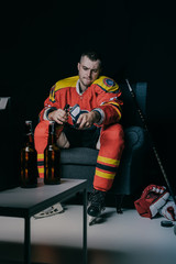 young hockey player using remote controller and drinking beer while sitting in armchair and watching tv on black
