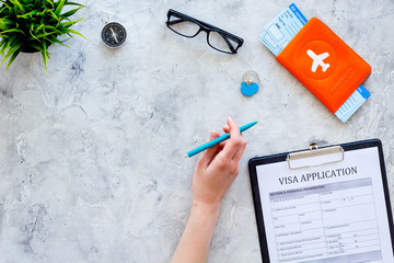Hand fills visa application form. Form near glasses, pen, passport cover with airplane signand airplane tiskets on grey background top view copy space