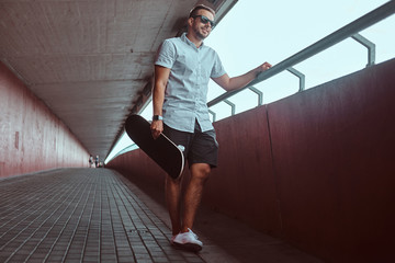 Smiling handsome fashionable skater guy in sunglasses dressed in a white shirt and shorts, leaning against a wall while standing on a footpath under the bridge, looking away.