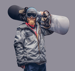 Portrait of a snowboarder dressed in a full protective gear for extream snowboarding posing with a snowboard on his shoulder at a studio. Isolated on gray background.