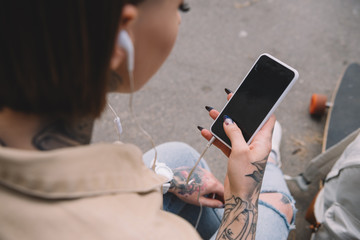 selective focus of tattooed woman in earphones listening music and holding smartphone with blank screen