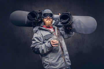 Redhead snowboarder with a full beard in a winter hat and protective glasses dressed in a snowboarding coat holds a snowboard and a bottle of alcohol on his shoulder in a studio.