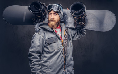 Brutal redhead snowboarder with a full beard in a winter hat and protective glasses dressed in a snowboarding coat holds a snowboard on his shoulder in a studio. Isolated on a gray background.