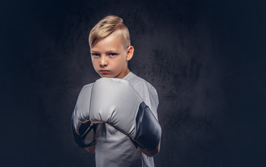 A handsome little boy boxer with blonde hair dressed in a white t-shirt in gloves ready to fight. Isolated on a dark textured background.