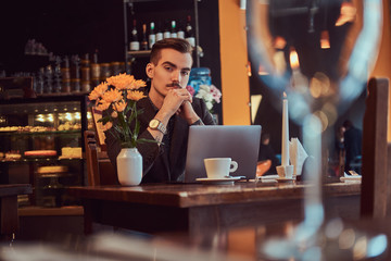 Portrait of a pensive handsome businessman with stylish beard and hair dressed in a black suit sitting with hand on chin at a cafe with an open laptop, lookong at the camera.