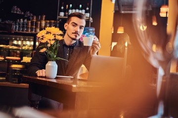 Handsome freelancer man with stylish beard and hair dressed in a black suit sitting at a cafe with an open laptop and holds a cup of coffee, looking at the camera.