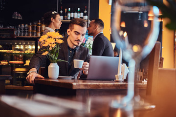 Handsome freelancer man with stylish beard and hair dressed in a black suit working on laptop while sitting at a cafe.