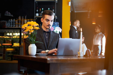 Handsome freelancer man with stylish beard and hair dressed in a black suit working on laptop while sitting at a cafe.
