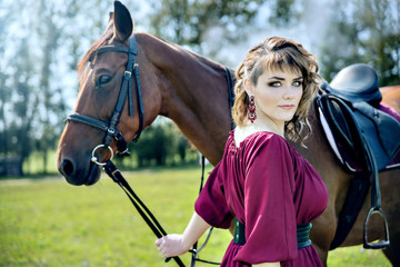 Obraz premium A beautiful girl in a burgundy dress is holding a brown horse and looking into the frame.