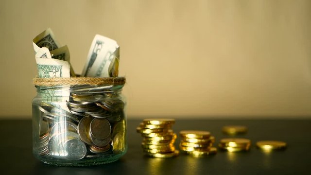Saving money coin in jar. Symbol of investing, keeping money concept. Collecting cash banknotes in glass tin as moneybox. American dollars close-up with increasing columns of gold coins on black table