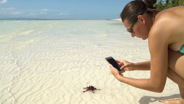 A woman in a swimsuit takes pictures of a starfish on a smartphone