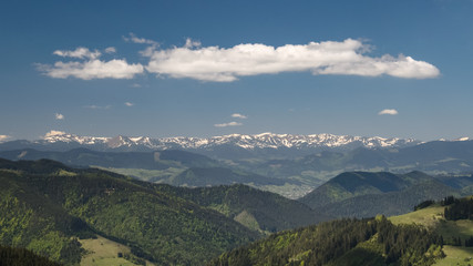 Scenic panoramic shot taken at sunny spring day. Mountains covered with forest and meadows, snowy mountains and blue sky with clouds. Concept of clear environment. Natural background.