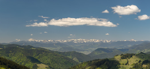 Scenic panoramic shot taken at sunny spring day. Mountains covered with forest and meadows, snowy mountains and blue sky with clouds. Concept of clear environment. Natural background.