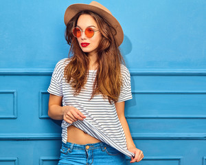 Young stylish girl model in casual summer clothes and brown hat with red lips, posing near blue wall
