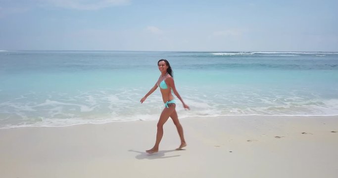 Camera moving parallel with beautiful woman running on white sand beach with blue ocean waves. Surfing school footage. Summer beach background. Sand and sea. Aerial drone slow motion footage Uhd 4K.
