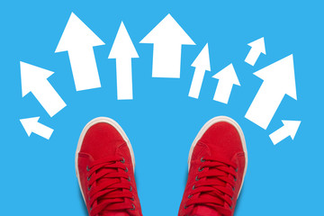 Legs in red sneakers on a blue background and a lot of arrows in different directions. Concept of many opportunities and freedom of choice. Flat lay, top view © Alex