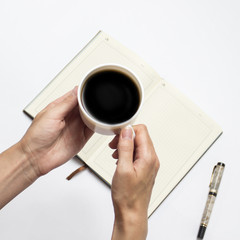Female hand with a cup of black coffee and an open diary with clean pages, a pen on a light background. Flat lay, top view