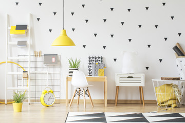 Yellow clock next to white chair at wooden desk in geometric kid's room interior. Real photo