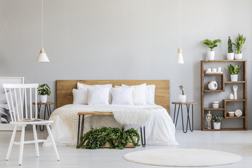 White chair near bed with wooden headboard in bright bedroom interior with plants. Real photo - obrazy, fototapety, plakaty