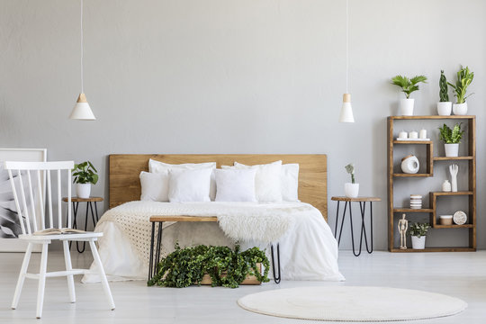 White chair near bed with wooden headboard in bright bedroom interior with plants. Real photo