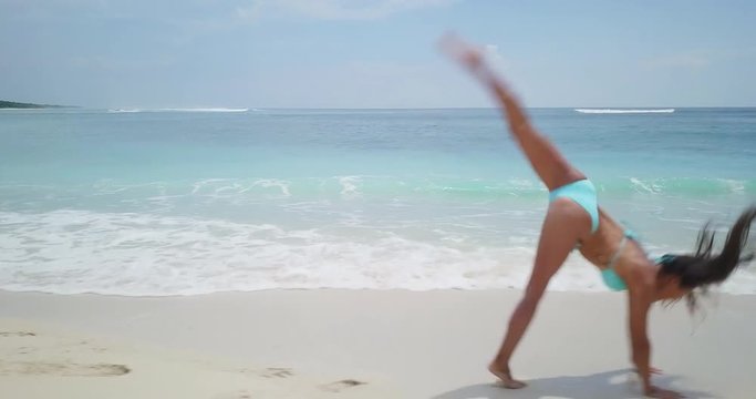 Camera moving parallel with beautiful lady doing gymnastic moves on white sand beach with blue ocean waves. Surfing school footage. Summer beach background. Aerial drone slow motion footage Uhd 4K.