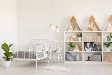 Plant and white chair next to bed in kid's bedroom interior with lamps and triangles. Real photo