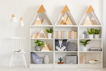 Lamps above white chair next to shelves with wooden triangles and plants in bright interior. Real...