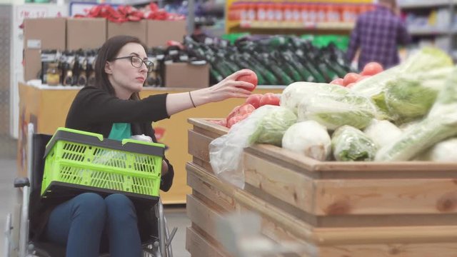Disabled woman in a wheelchair shopping in the supermarket chooses tomatoes and puts them in a package.