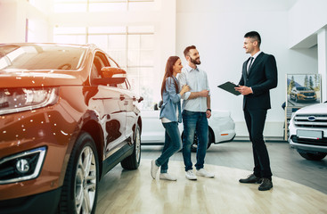 Young modern happy couple buying a new car in city dealership and talking with salesman - obrazy, fototapety, plakaty