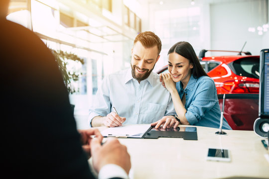 Beautiful happy couple sitting at the table signing a documents for the purchase of a car at the dealership