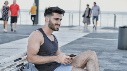 Young handsome man using smartphone, typing text message to someone while sitting on a bench at the seaside over countryside landscape