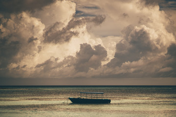 A lonely boat is lying at anchor in the open sea during the evening with a stunning cloudscape above on the sunset  an empty anchored blue vessel in the ocean not far from the coast, seascape behind © skyNext