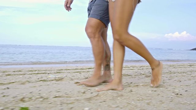 Low section of tanned legs of boyfriend and girlfriend walking barefoot on sandy ocean beach