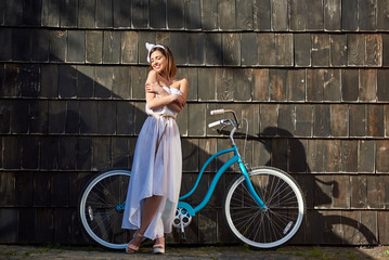 Horizontal full length shot of a joyful young attractive woman embracing herself smiling with her eyes closed standing near her bicycle outdoors on a warm sunny day.