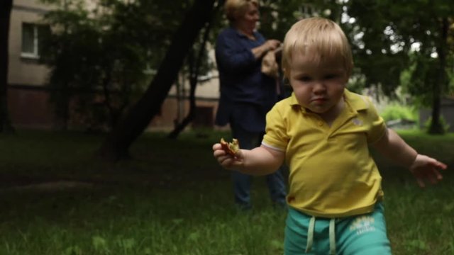 happy cute smiling blonde one year old baby boy walking himself on a grass in beautiful sunny summer day with grandmother using smartphone mobile on blur background internet addiction children child