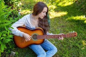 Young hipster woman sitting in grass and playing guitar on park or garden background. Teen girl...