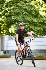 Fototapeta na wymiar Young male bicyclist in cycling sportswear and protective helmet, looking at camera, riding bike down empty city center alley surrounded by green trees. Sportsman training outdoors improving skills.