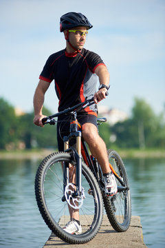 Young man biker in professional cycling clothing and helmet posing near bike looking in distance, thinking about future success and achievement. Sportsman taking break at pier near lake.