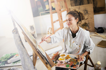 Artistic girl sitting in studio and paint on easel.