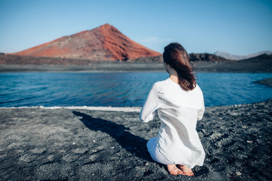 Back view of woman in white clothes praying in black sand beach