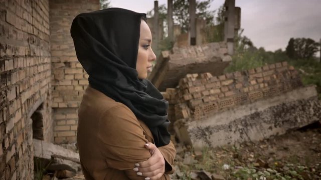 Young muslim woman in hijab standing near ruined building and looking at camera with scared and worried expression, ruin in background