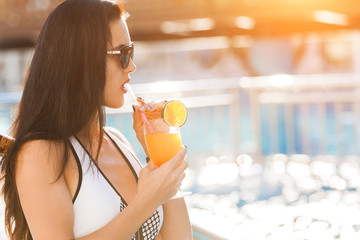 Pretty slim woman with long dark hair standing near the swimming pool with a cocktail wearing white stylish swimwear and sunglasses. concept of luxury holiday time. free copyspace