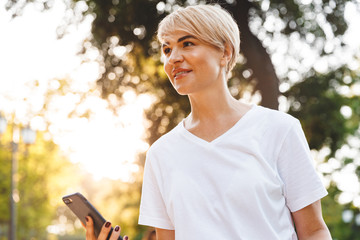 Photo of happy caucasian woman wearing casual clothing smiling, while holding smartphone during walk in green city park