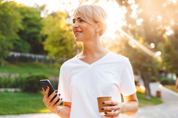 Image of beautiful happy woman with blond hair wearing casual clothing holding cell phone and takeaway coffee, during walk in green park