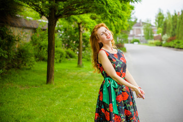 Fototapeta premium Retro girl with red lips in a dress with a print of roses on a beautiful summer background. Young redhead model on a background of a bush of roses. Stylish woman. Fashionable summer dress 