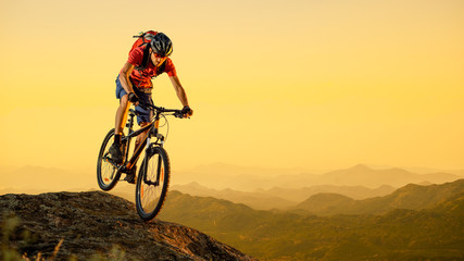 Fototapeta premium Cyclist in Red Riding the Bike Down the Rock at Sunset. Extreme Sport and Enduro Biking Concept.