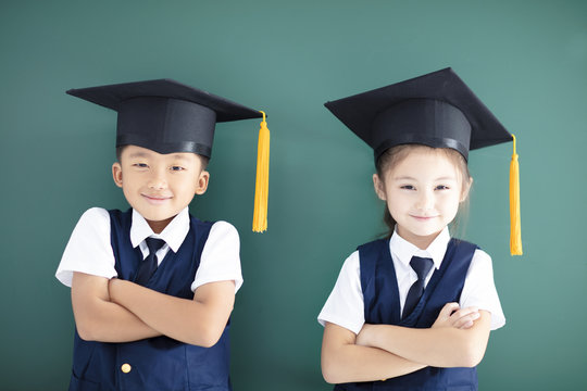 Happy boy and girl in graduation cap stand before chalkboard