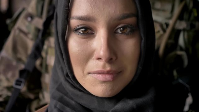 Close portrait of young muslim woman in hijab crying and looking at camera, armed soldier with weapon standing behind woman, war