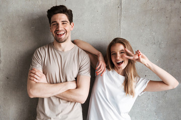 Photo of joyful caucasian couple girl and guy in basic clothing posing together at camera with happy smile and peace sign, isolated over concrete gray wall indoor
